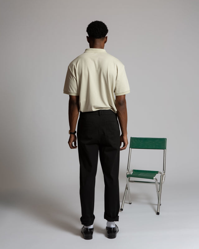 Back view of man wearing Kadet men's performance polo shirt in beige with black pants standing near green chair