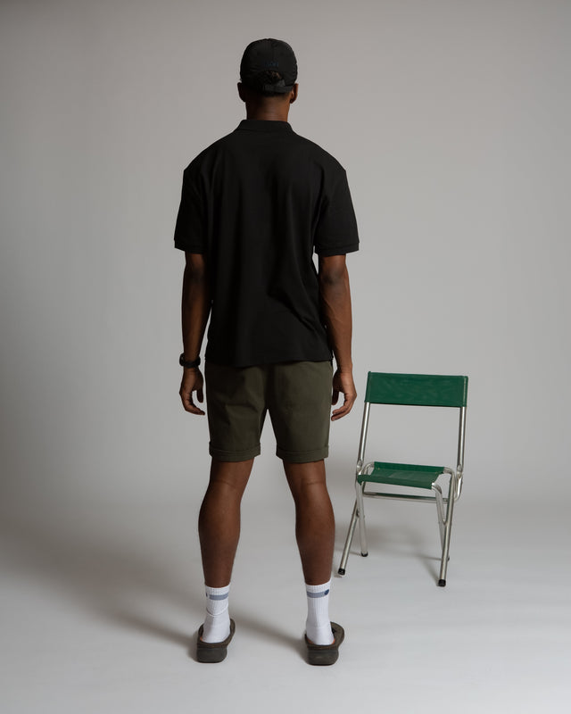Back view of a man wearing a black men's performance polo shirt by Kadet with olive shorts and black cap