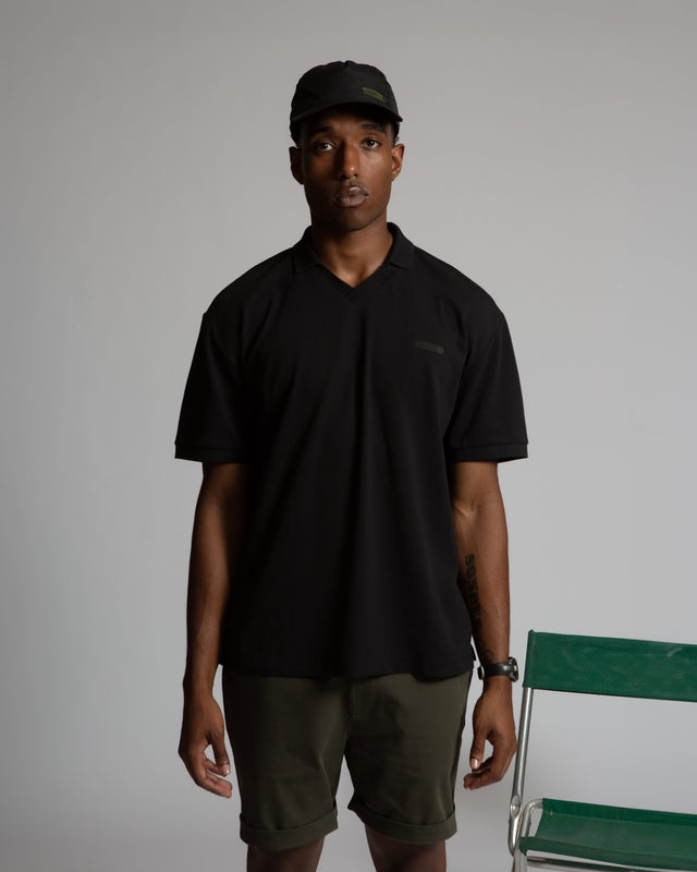 Kadet men's performance polo shirt in black worn with shorts and a cap in a studio setting