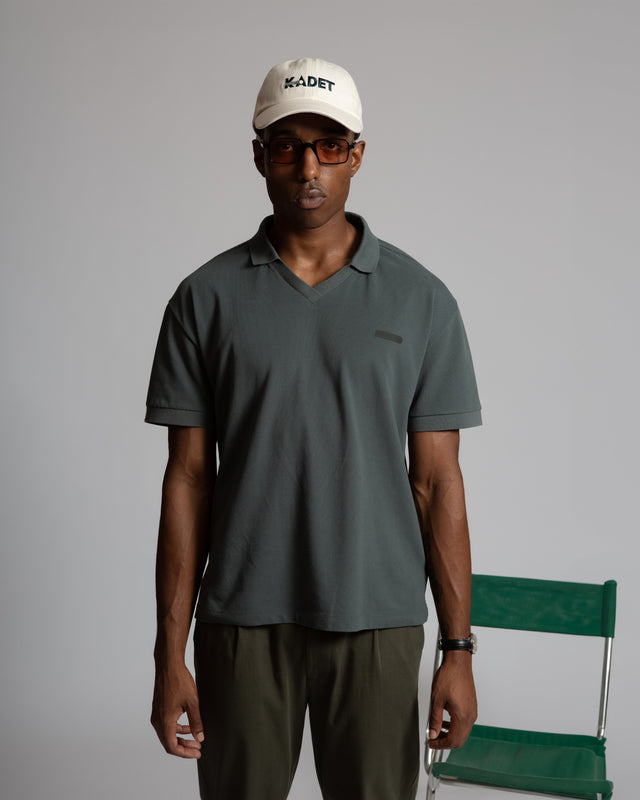 Kadet men's performance polo shirt in dark gray worn by a male model with glasses and a white Kadet cap