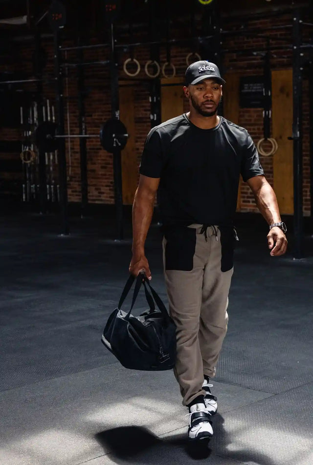 Man wearing Solstice Athletics premium workout clothing with black t-shirt and gray pants carrying a gym bag