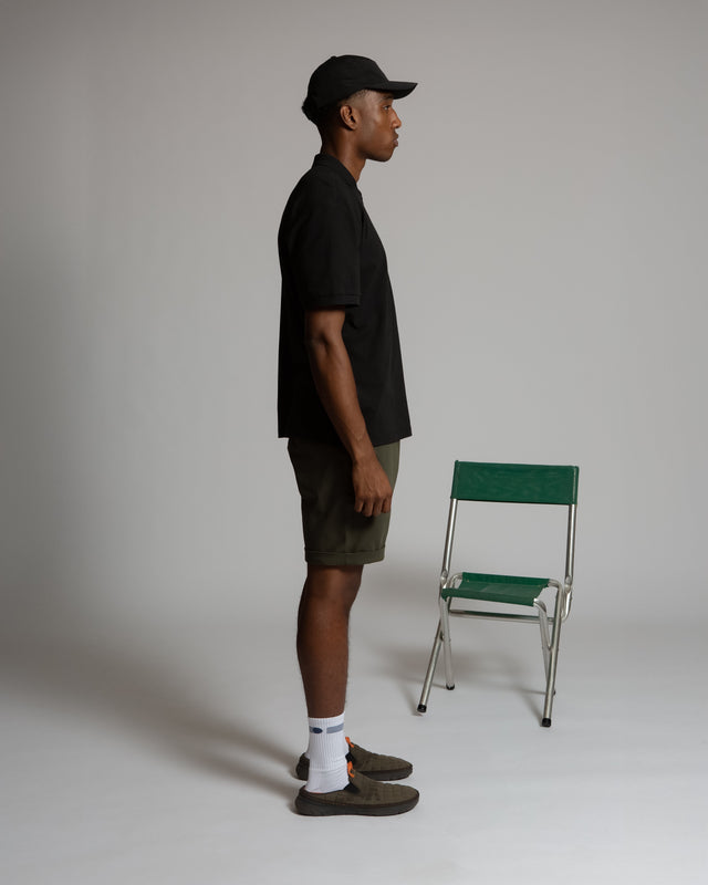 Side view of a man wearing black shirt and olive green men's chino shorts with white socks and black cap
