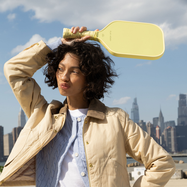 Woman holding yellow Play Henry carbon fiber pickleball paddle outdoors with city skyline background