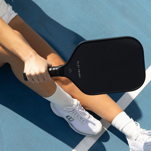 Play Henry black carbon fiber pickleball paddle held by player sitting on court in white shoes and socks