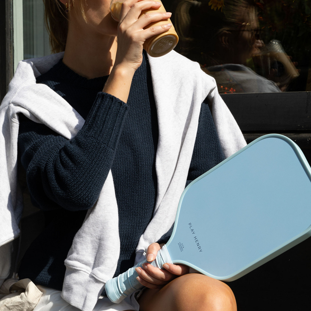 Play Henry blue pickleball paddle held by person wearing navy sweater and white hoodie drinking coffee