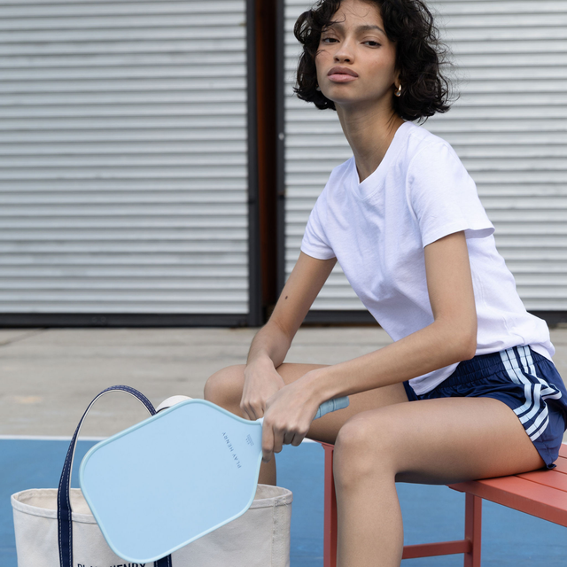 Woman sitting on bench holding light blue Play Henry pickleball paddle outdoors on court