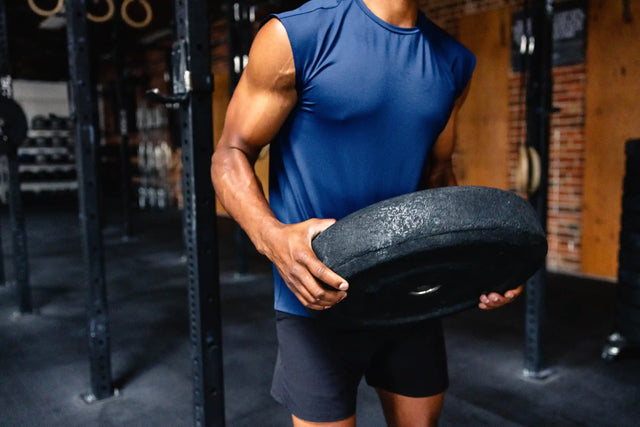 Man wearing Solstice Athletics premium workout clothing sleeveless blue shirt and black shorts holding weight plate